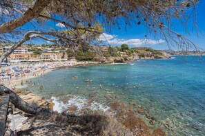 Ein Strand mit klarem Wasser, felsigem Ufer und Gebäuden im Hintergrund.