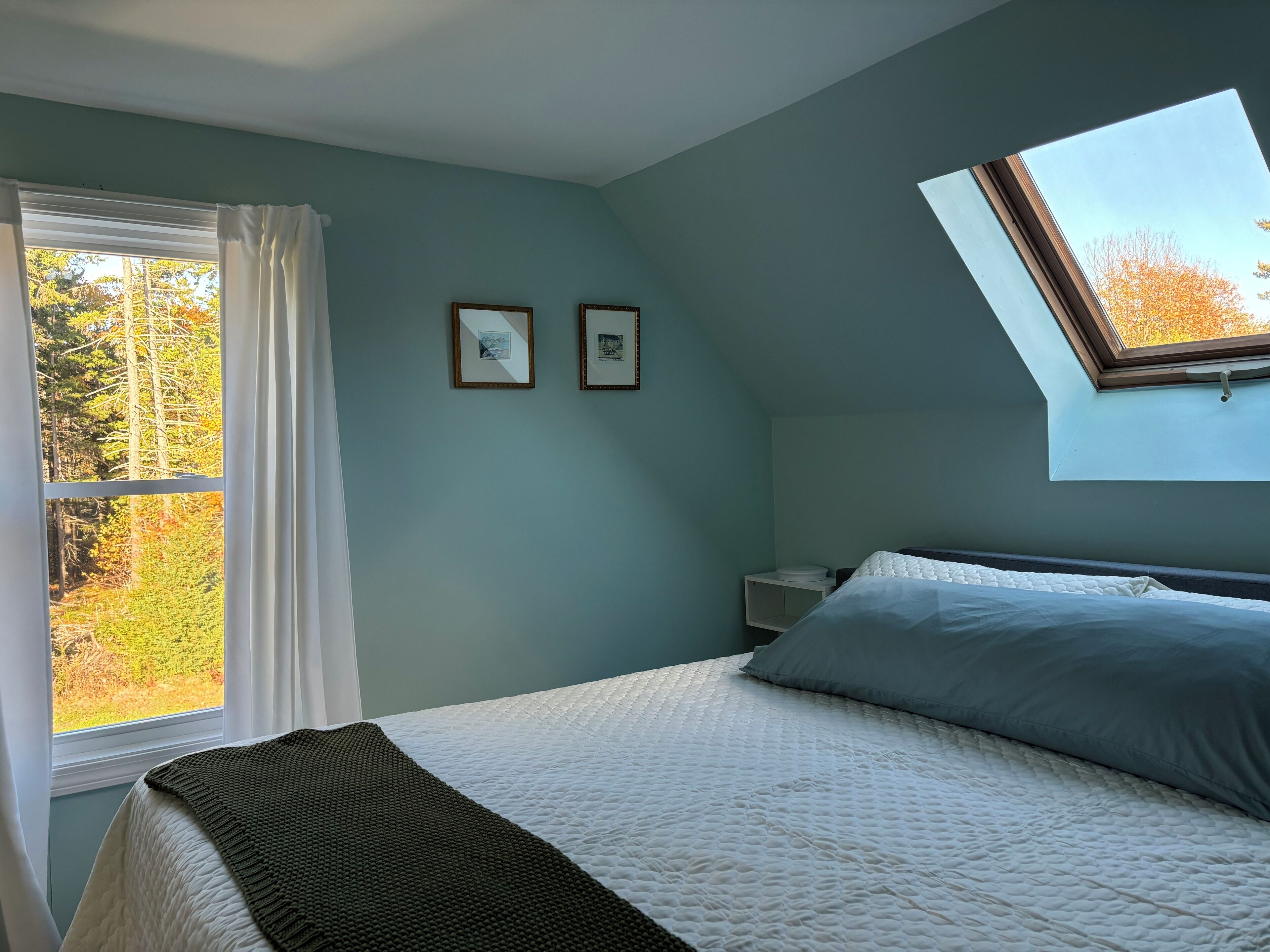 A bedroom with a bed, a skylight, a window with curtains, and two framed pictures on the wall.
