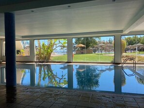 Une piscine intérieure avec des parois en verre transparent, un sol carrelé et une vue sur un espace extérieur agrémenté de parasols et de verdure.