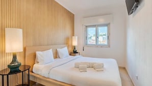 Une chambre d’hôtel avec une tête de lit en bois, un grand lit avec du linge de lit blanc, deux lampes de chevet, un téléviseur à écran plat fixé au mur et une fenêtre donnant sur la ville.