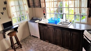 Una cocina compacta con estufa, lavabo y lavadora. Hay dos ventanas con cortinas, una laptop sobre una mesita y un escurreplatos azul.