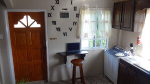Una cocina con puerta de madera, una laptop sobre una mesita, una ventana con cortinas y un lavamanos con armarios.