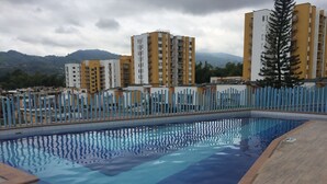 Une piscine avec une terrasse carrelée, entourée d’une clôture, et des bâtiments en arrière-plan.