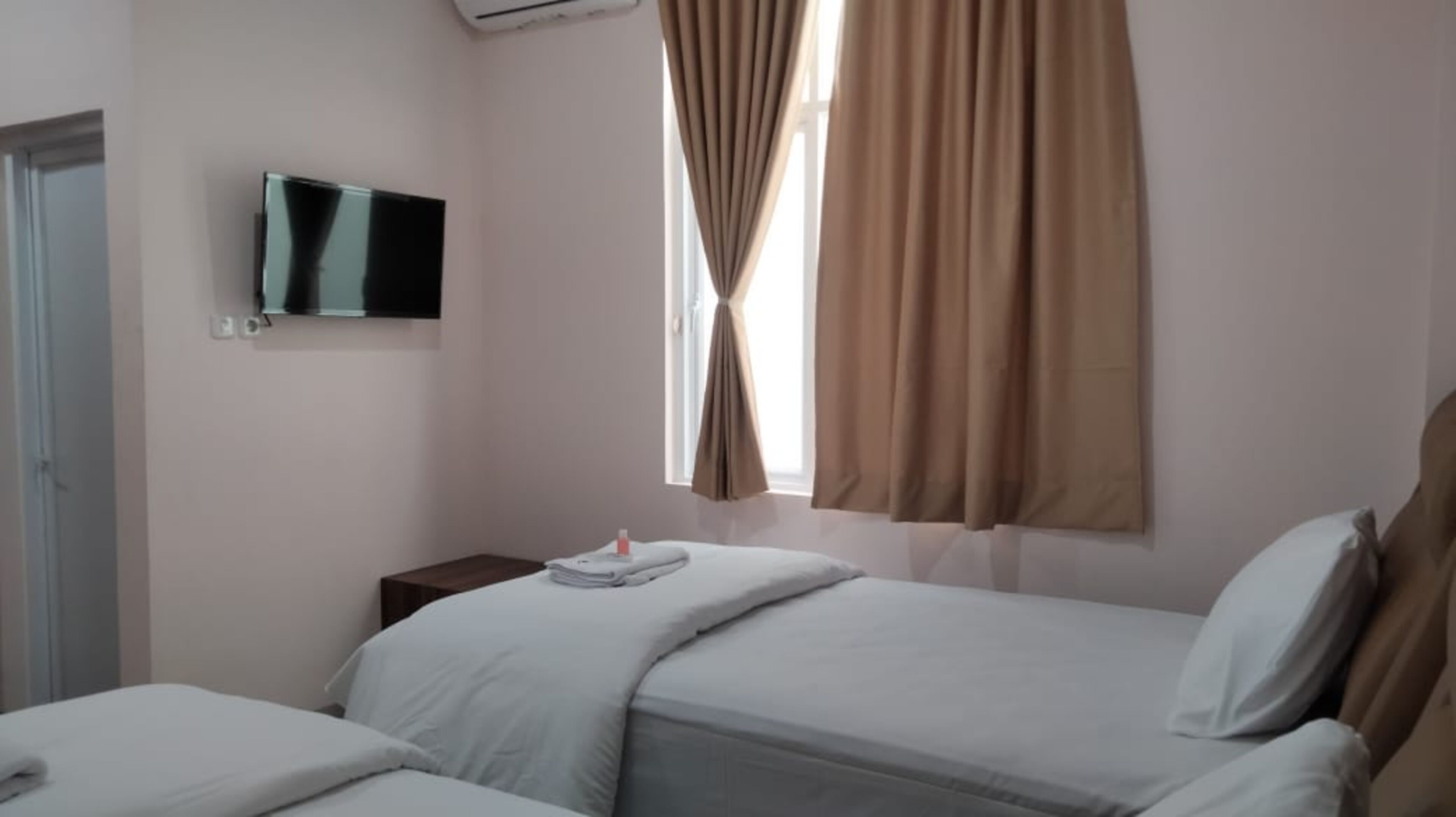 A hotel room with a bed, a small bedside table with a bottle, a TV mounted on the wall, and a window with curtains.