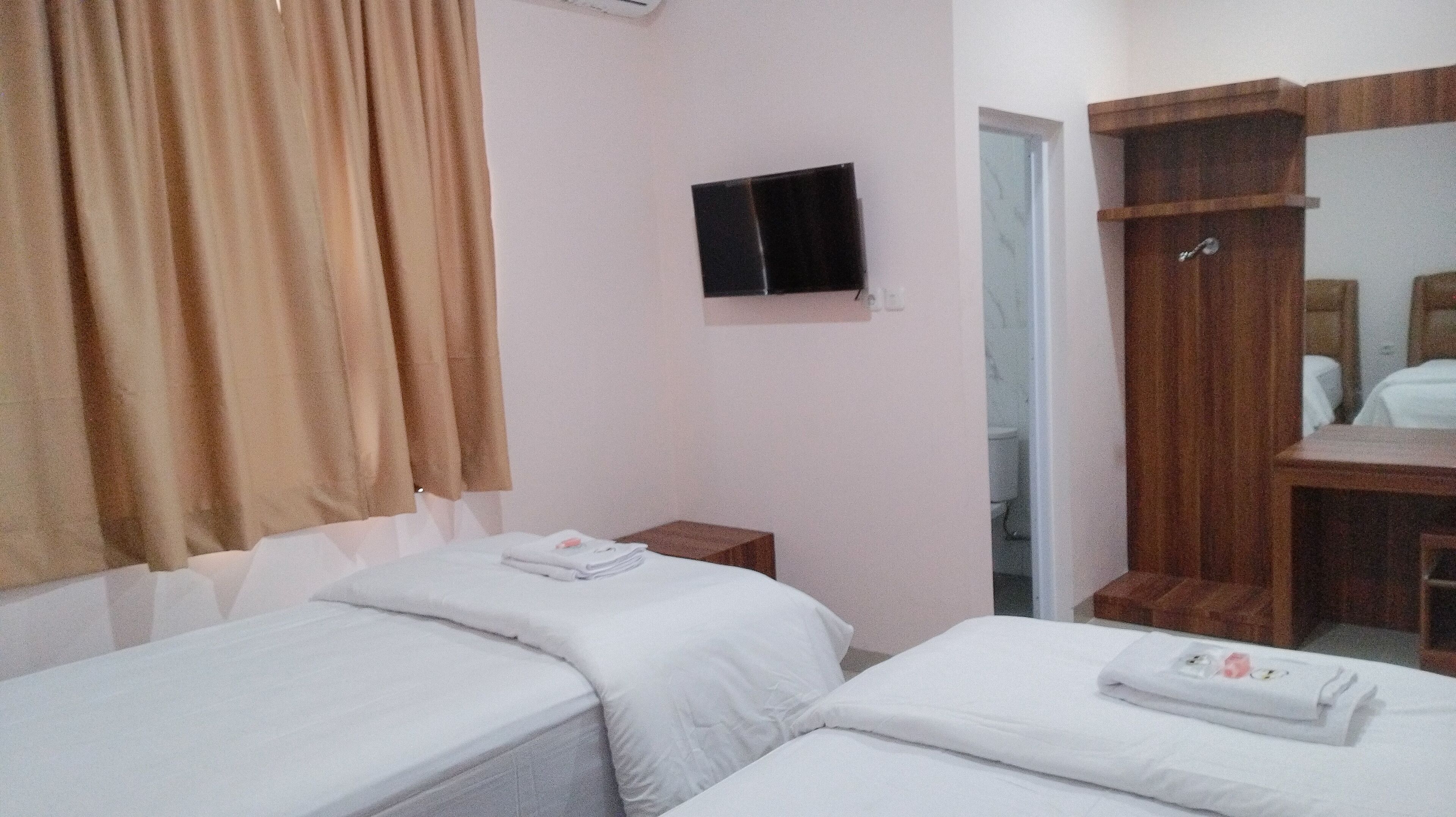 A hotel room with two single beds, a wooden wardrobe, a television mounted on the wall, and a bathroom visible through an open door.