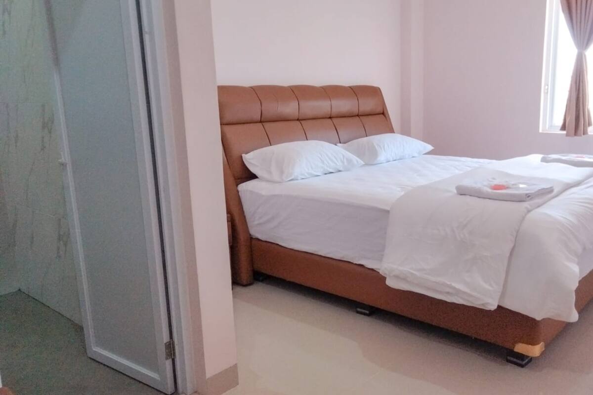 A neatly made bed with white linens and a brown headboard in a room with a plain white wall and a window with curtains.