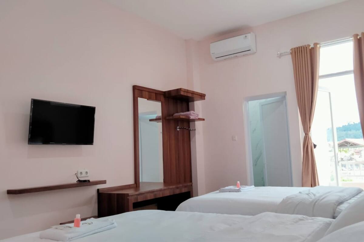 A hotel room with two beds, a TV, a mirror, an air conditioner, and a window with curtains.
