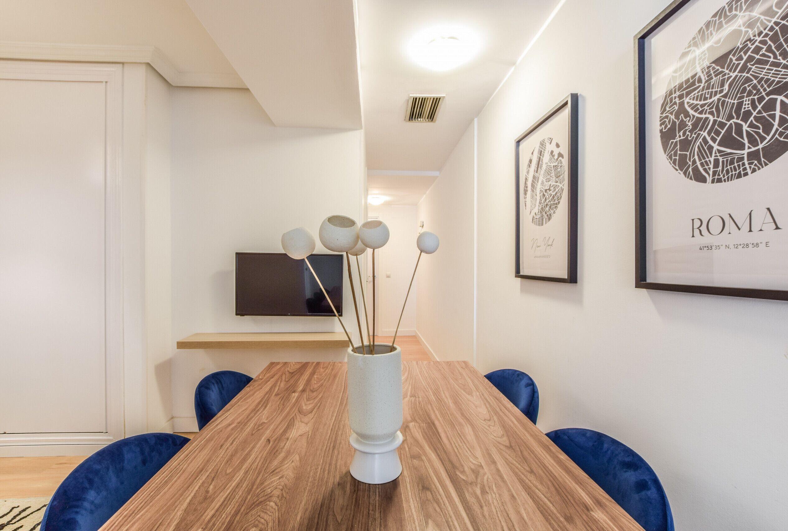 A modern dining area with a wooden table, blue chairs, a white vase with spherical decorations, and framed artwork on the walls.