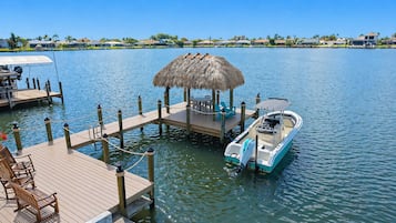 A dock with a boat, a thatched-roof structure, and wooden chairs.