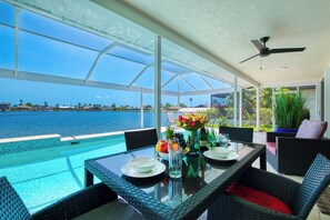 A dining area with a view of a body of water, a glass table, wicker chairs, and a ceiling fan.