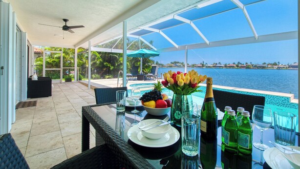 Un coin repas avec une table dressée pour deux personnes, donnant sur une piscine et un plan d’eau.