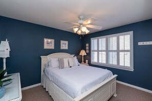Quarto com cama, ventoinha no teto, janela com persianas e mesa de cabeceira com abajur.