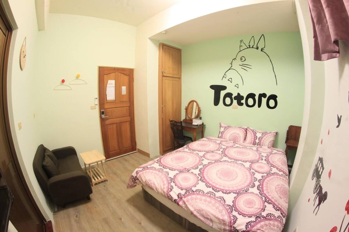 A bedroom with a bed, a small table, a chair, a wooden door, and a wall with a Totoro drawing.
