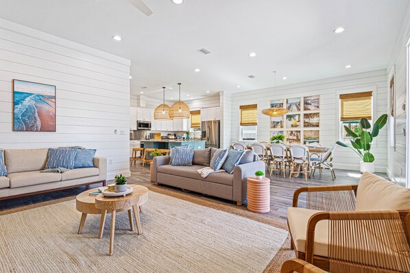 A spacious living area with a sofa, coffee table, and wicker chairs. A dining area with a table and chairs is visible in the background.