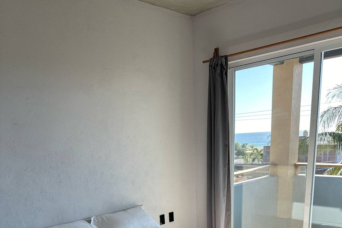Una cama bien tendida con sábanas y toallas blancas, un cabecero de madera y vistas al océano a través de un ventanal con cortina.