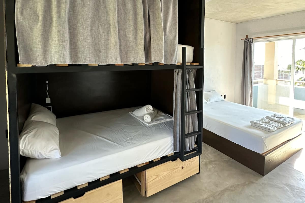 Una litera con estructura de madera, una cama individual abajo y una ventana con cortinas.