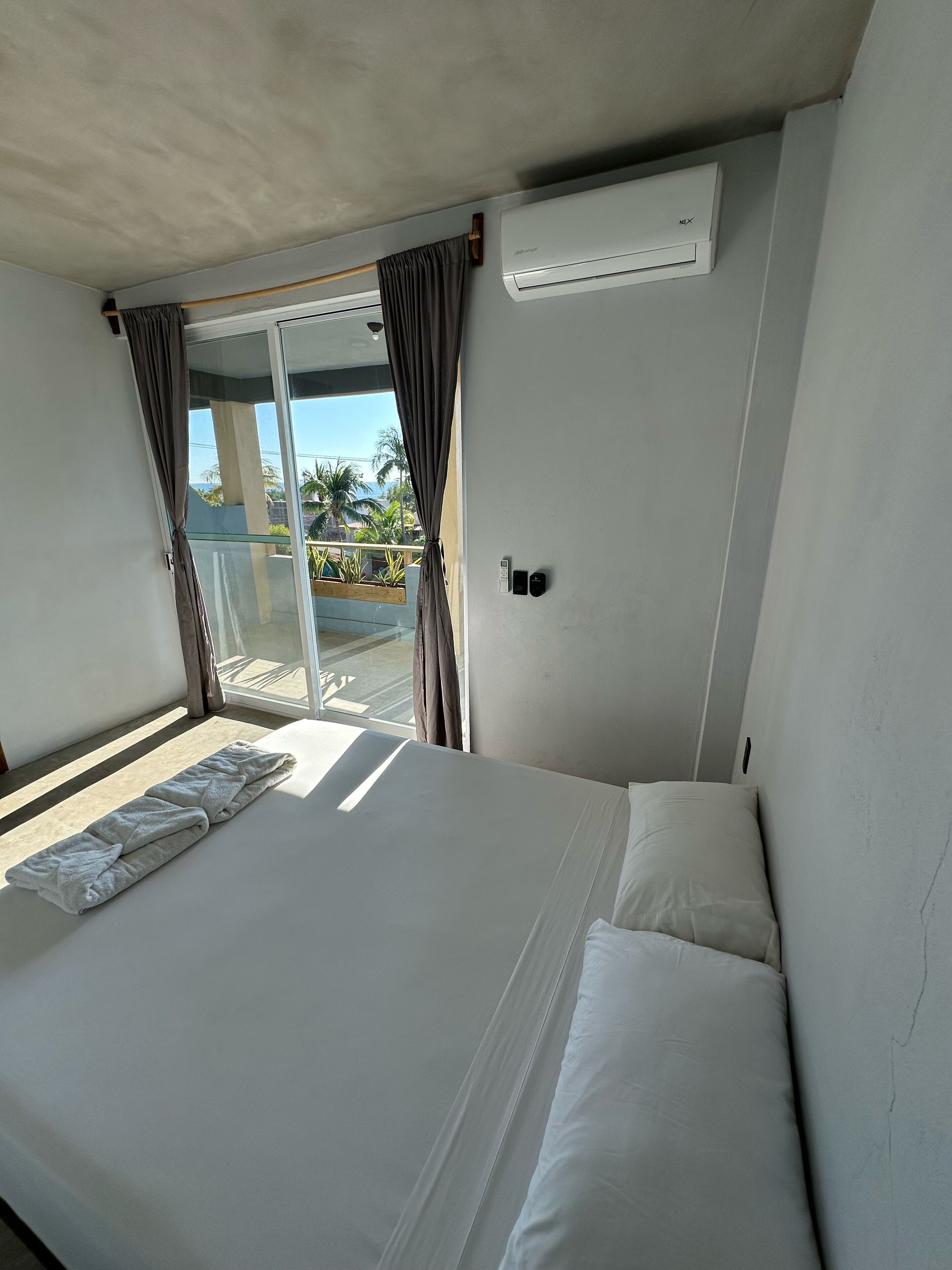 A hotel room with a bed, a window with curtains, an air conditioning unit, and a view of palm trees and buildings outside.