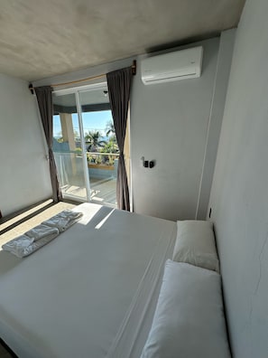 A hotel room with a bed, a window with curtains, an air conditioning unit, and a view of palm trees and buildings outside. - Straw Hat Surf Hostel (Puerto Escondido)