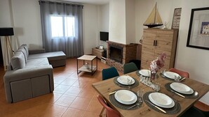 Ett modernt vardagsrum med en soffa, ett matbord för fyra personer, en öppen spis och en tv.