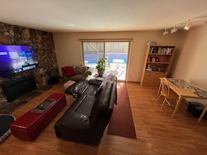 Un salon avec un mur en pierre, un canapé en cuir noir, un pouf rouge, un téléviseur diffusant des services de streaming, une table à manger en bois avec des chaises et une bibliothèque.