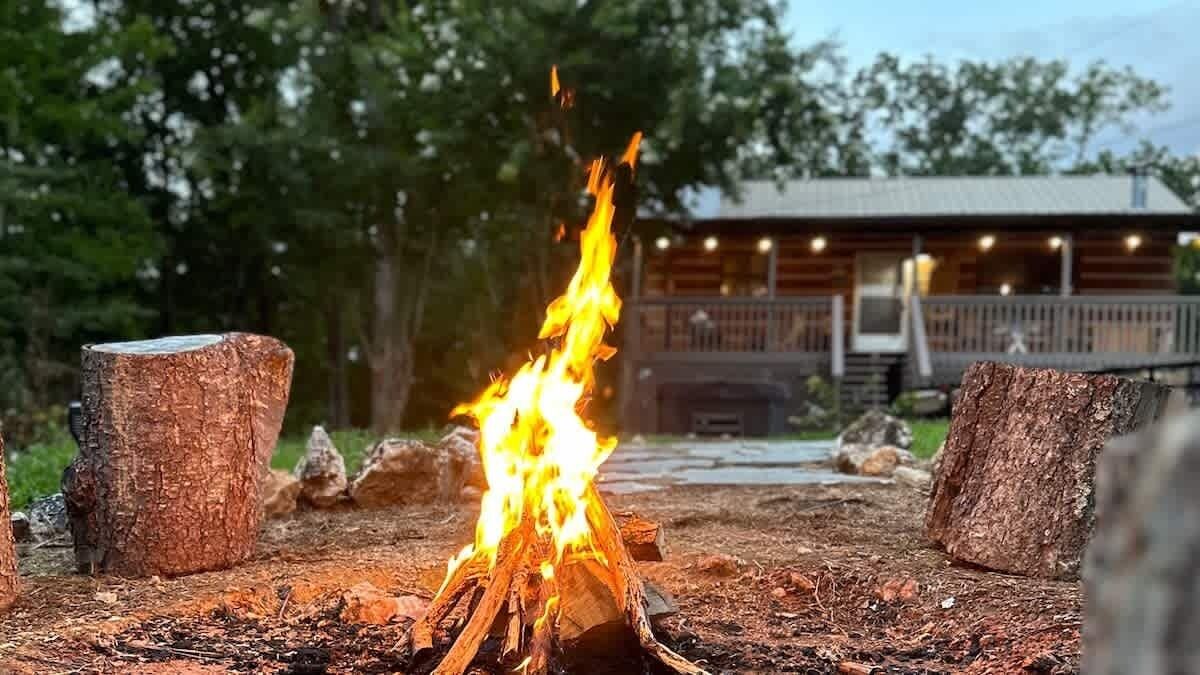 A campfire with logs and a wooden stump, surrounded by rocks, with a cabin in the background.
