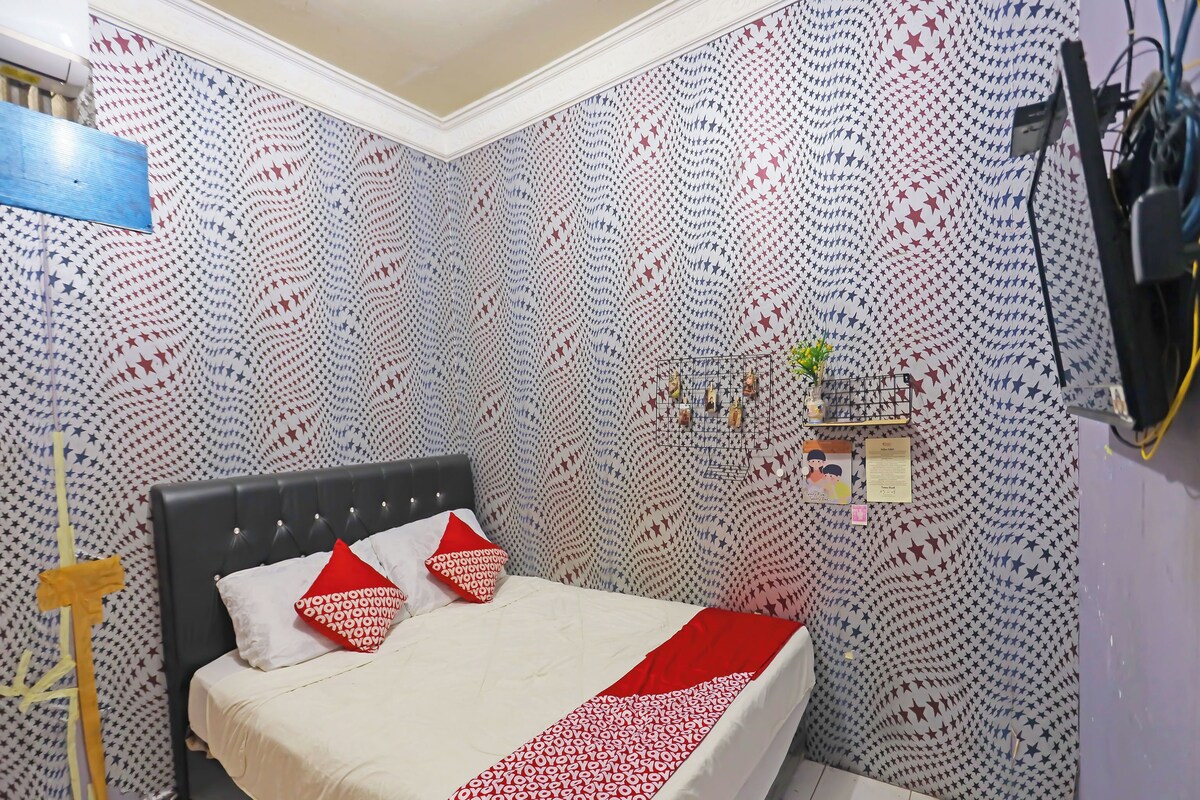 A room with a bed, a wall-mounted TV, and a small shelf with a plant and a card.