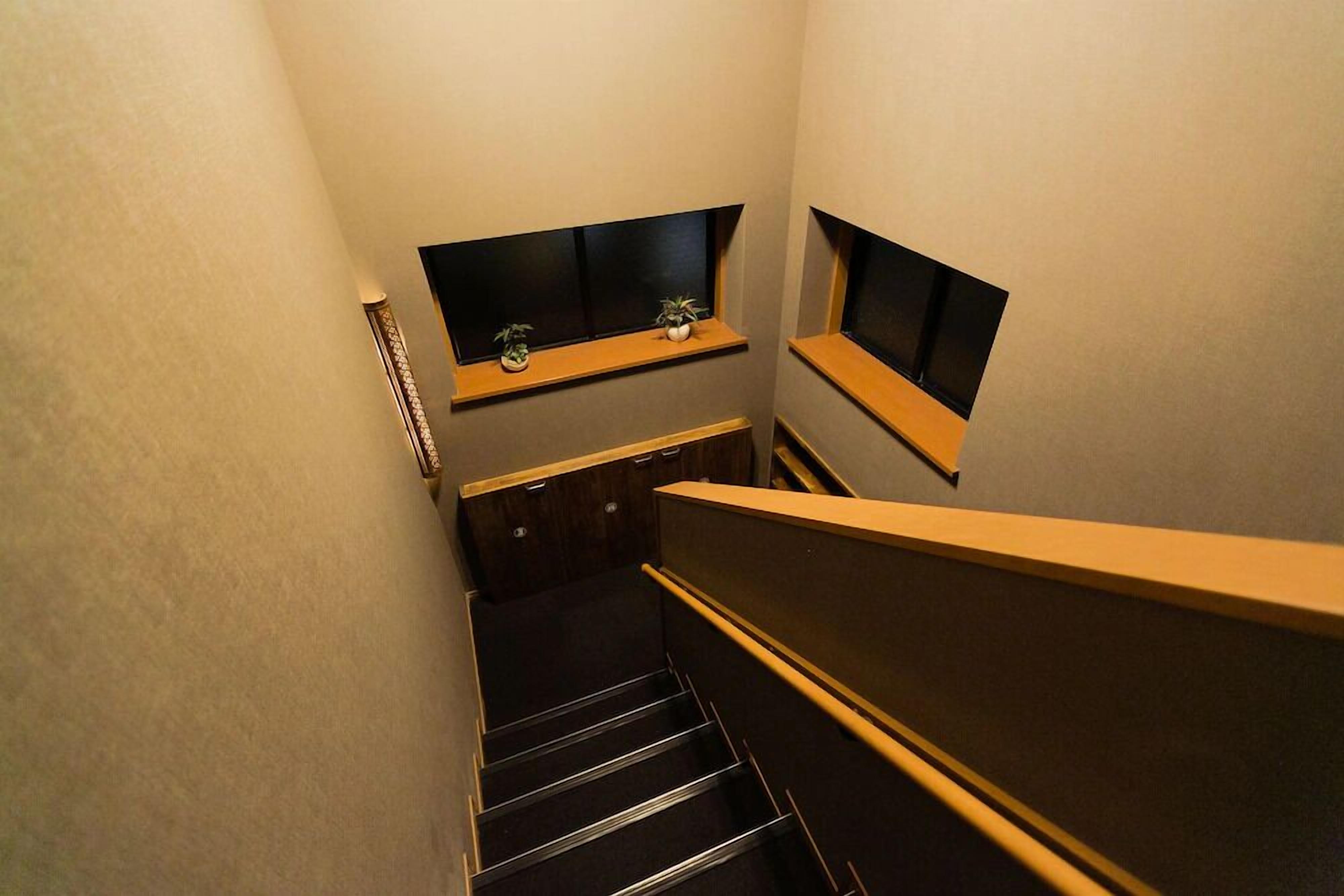 A narrow stairwell with wooden steps and a handrail, leading to a landing with two small windows and a shelf with a plant.