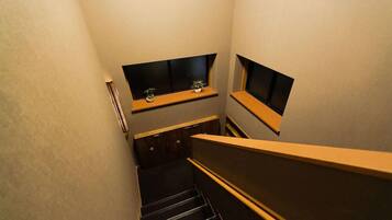A narrow stairwell with wooden steps and a handrail, leading to a landing with two small windows and a shelf with a plant.
