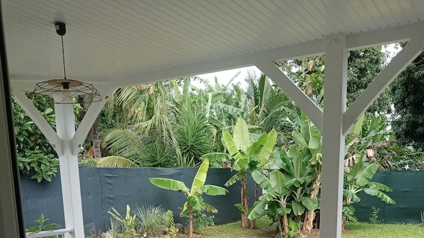 Uma área de varanda coberta com uma mesa e cadeiras acessíveis para cadeiras de rodas, cercada por um jardim exuberante com bananeiras e um cercado verde.