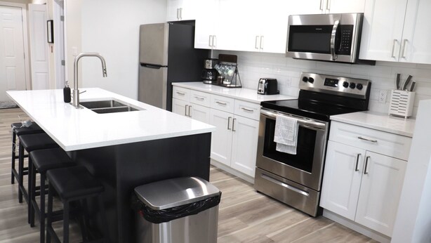 A modern kitchen with white cabinets, stainless steel appliances, a central island with bar stools, and a trash can. - Rare*Full Home in Downtown Winnipeg|Free Parking (Winnipeg)