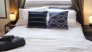 A neatly made bed with white linens and two patterned pillows, a window with curtains, and bedside lamps on a nightstand. - Rare*Full Home in Downtown Winnipeg|Free Parking (Winnipeg)