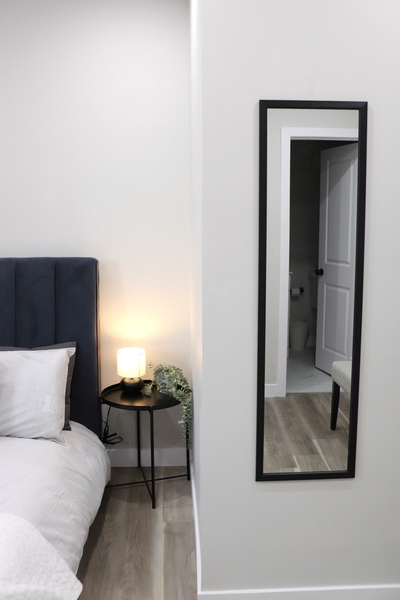A bedroom with a bed, bedside table with a lamp, a mirror, and a door leading to a bathroom.