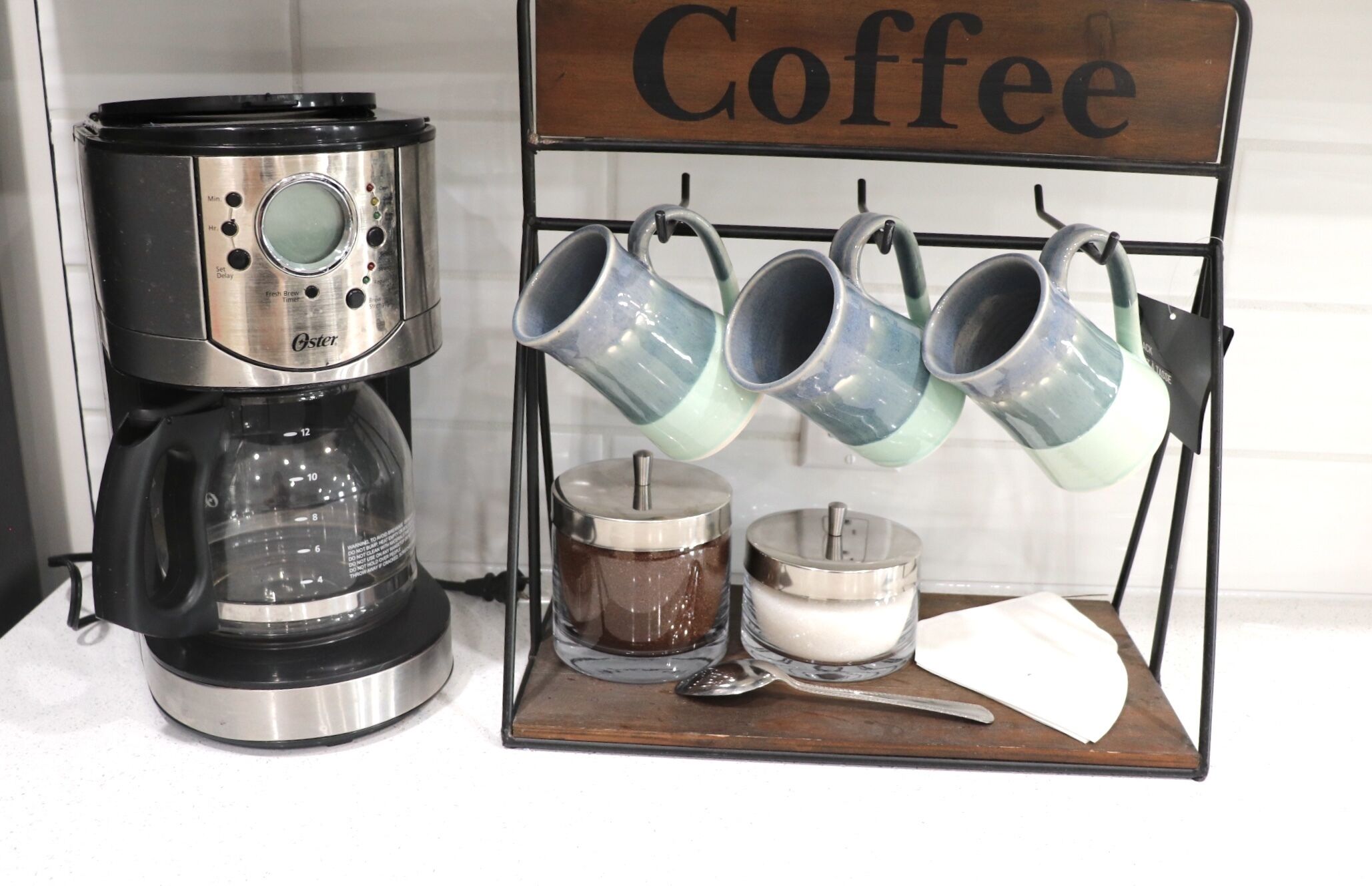 A coffee maker with a timer and a coffee mug rack with several mugs and a spoon on a wooden board.