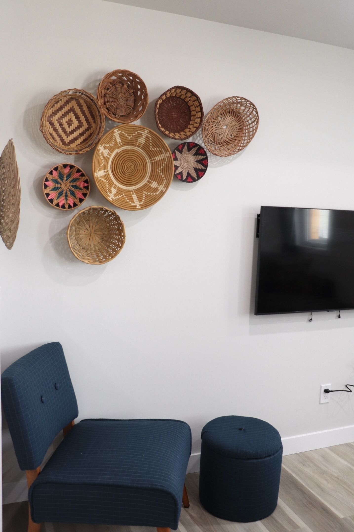 A living room with a flat-screen TV mounted on the wall, a blue armchair, a matching ottoman, and a collection of woven baskets on the wall.