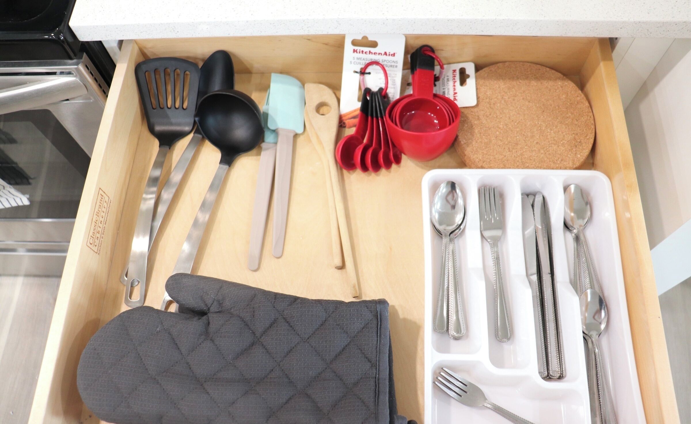A kitchen drawer with various utensils, including a black oven mitt, measuring spoons, a red measuring cup, and a white plate with cutlery.