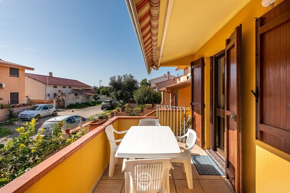 A balcony with a white table and chairs, a yellow wall, and a view of neighboring houses and parked cars.
