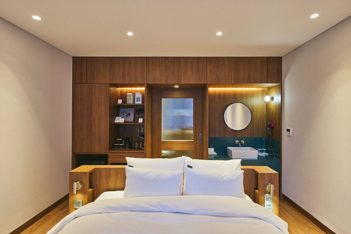 A modern hotel room with a wooden bed, a built-in shelf with toiletries, a round mirror, and a view through a frosted glass door.