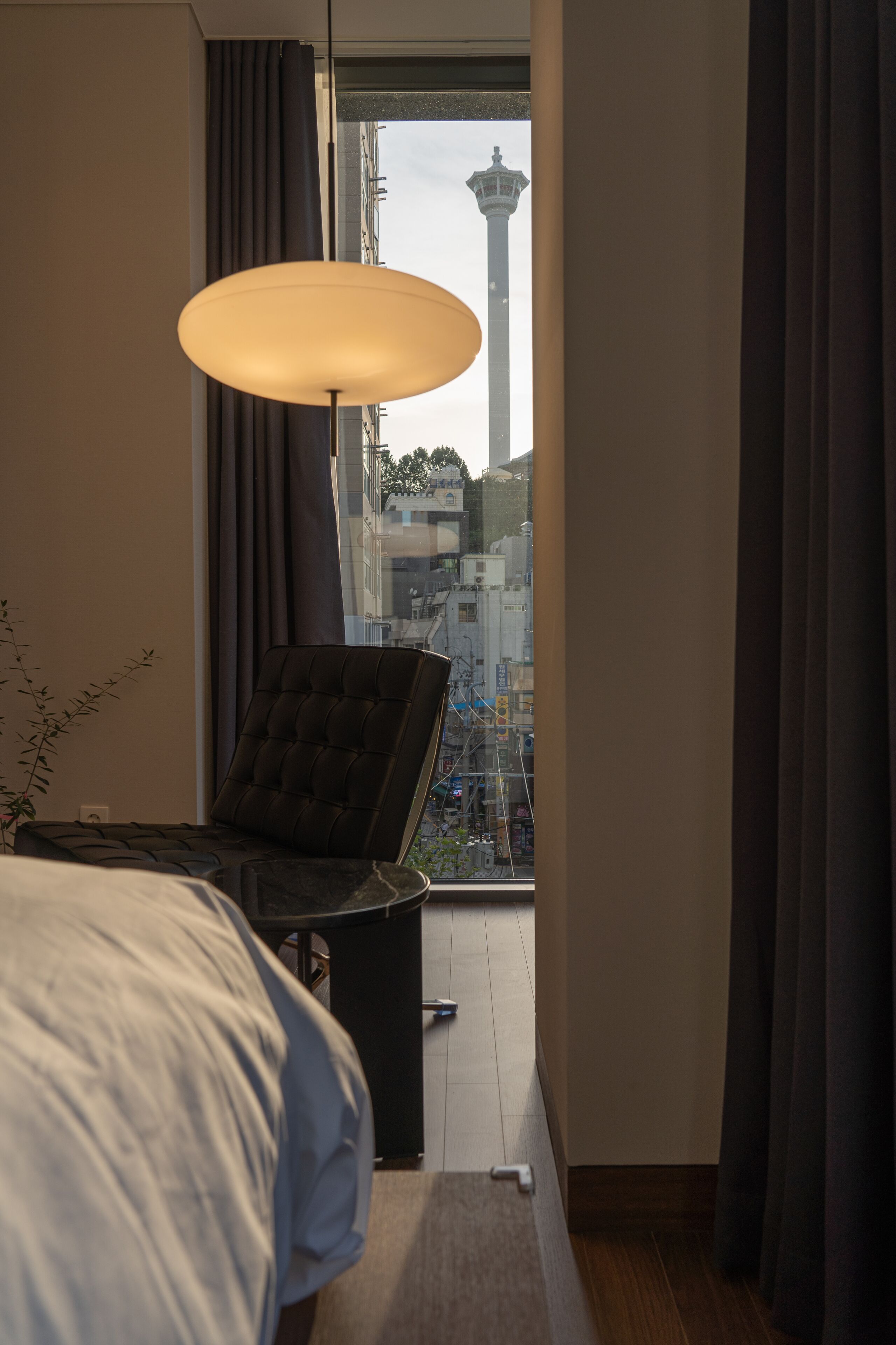 A hotel room with a bed, a chair, a desk, a lamp, and a view of a tower through a window.