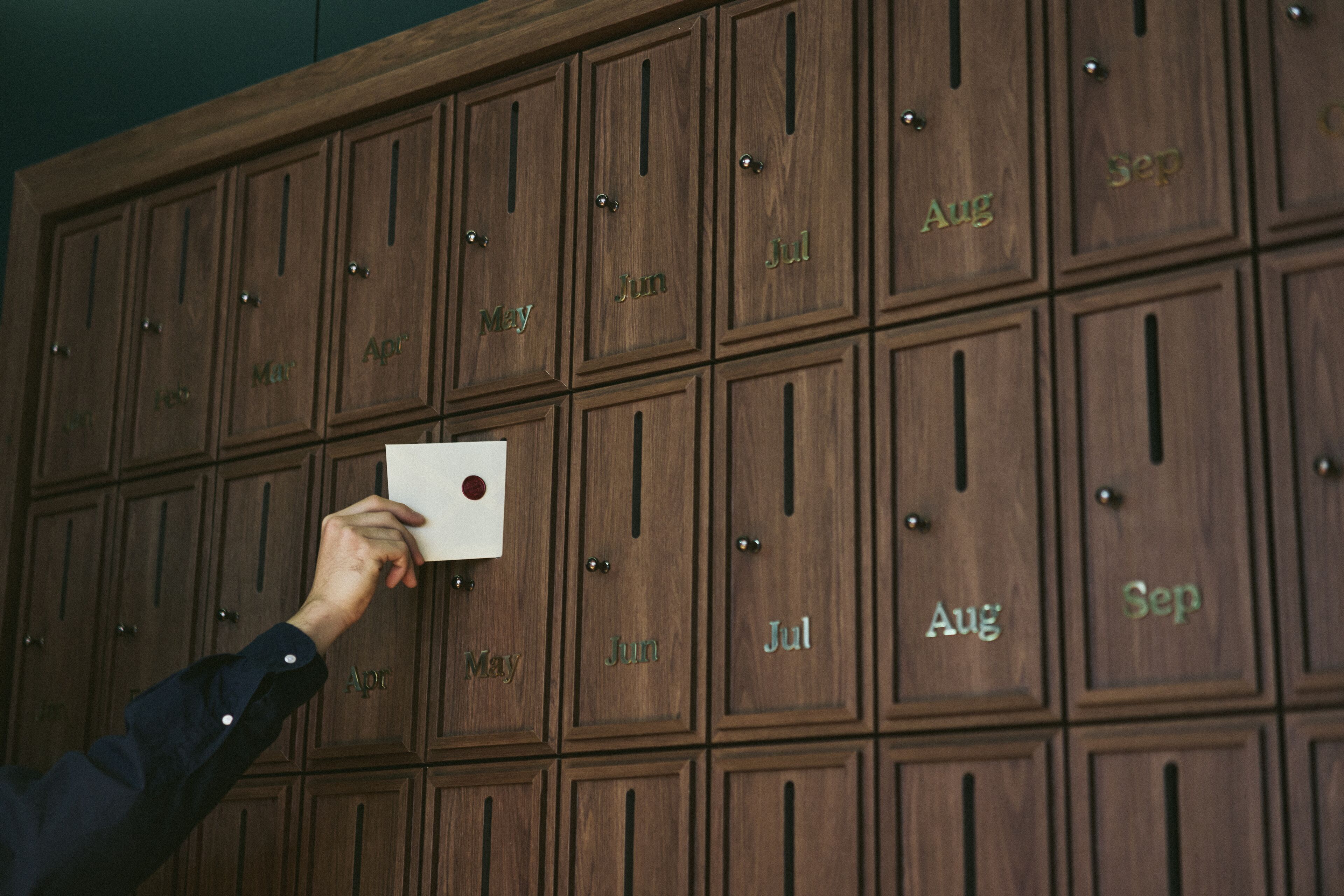 A hand holding a card in front of a wall of wooden lockers with months labeled on them.
