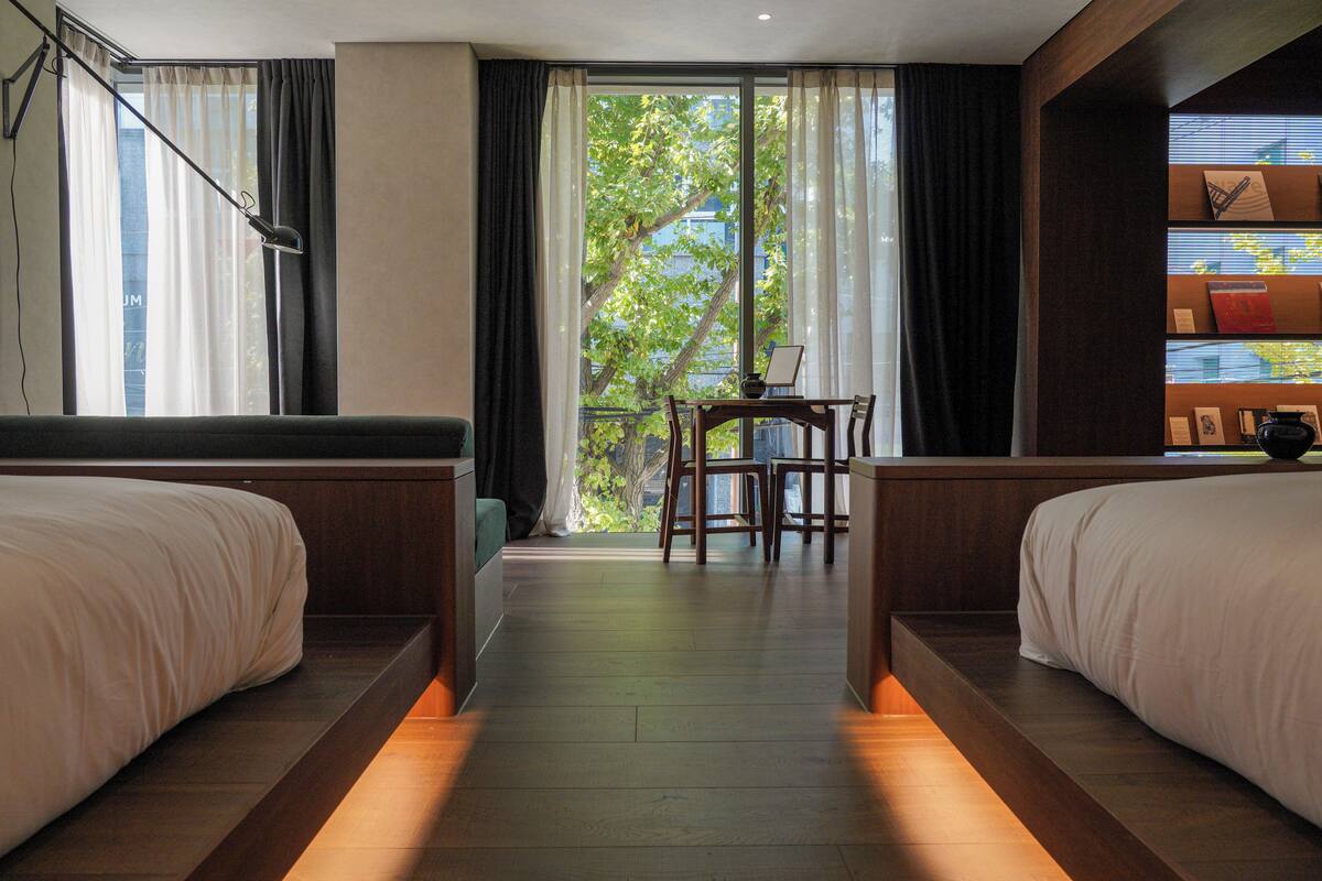 A hotel room with two beds, a small table, and a view of greenery through the window.