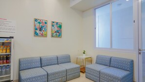 Ein Zimmer mit einem blauen, gemusterten Sofa, einem kleinen weißen Tisch mit einer Pflanze und zwei farbenfrohen Bildern an der Wand.