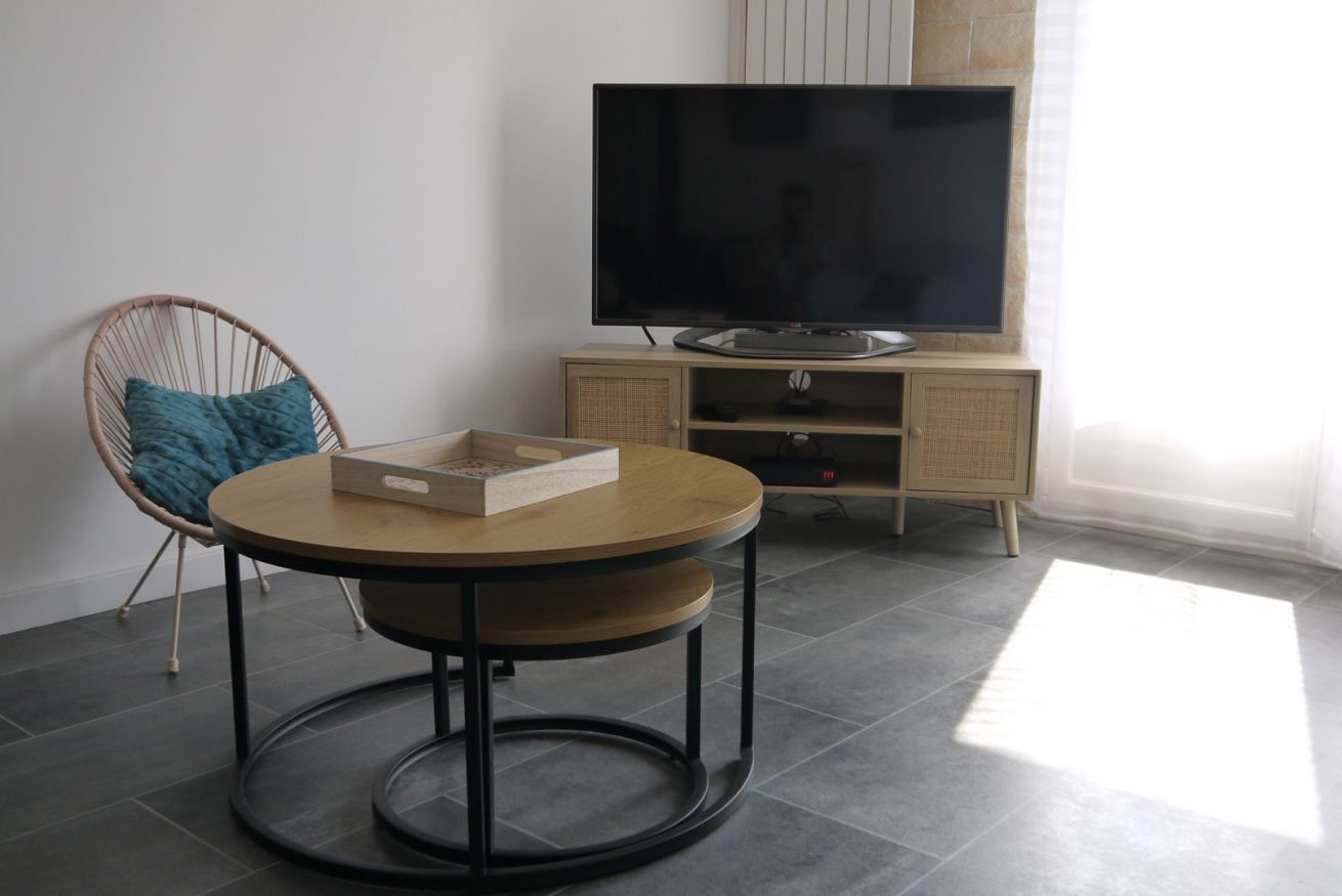 Une pièce avec une table ronde, une chaise en osier, un téléviseur à écran plat posé sur un meuble et un sol carrelé.