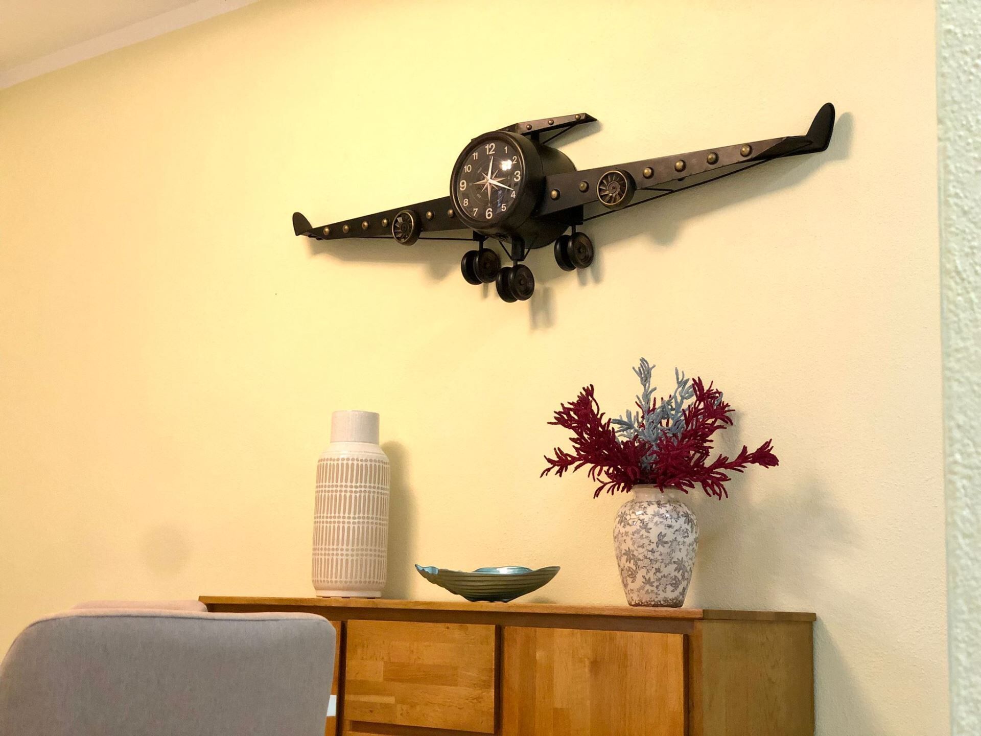 A wall-mounted clock with a unique design, a white bottle, a bowl, and a floral vase on a wooden cabinet.