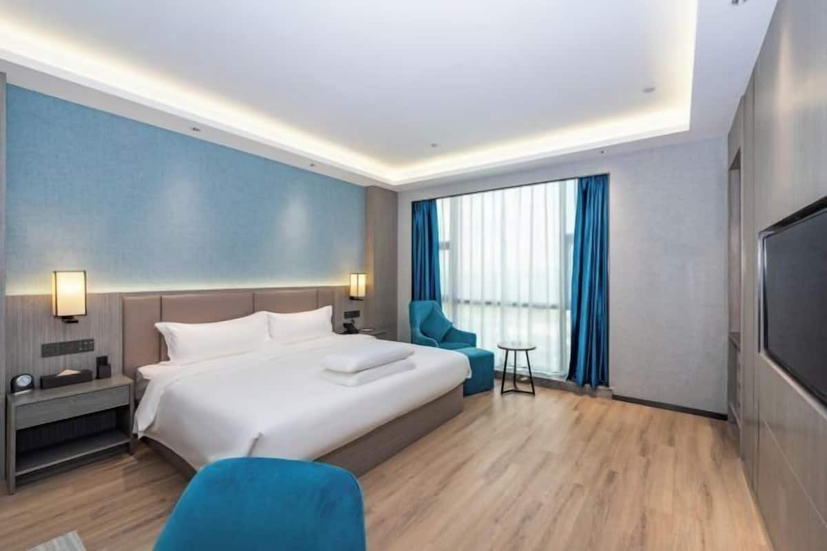 A modern hotel room with a large bed, a seating area, a TV, and a window with curtains.