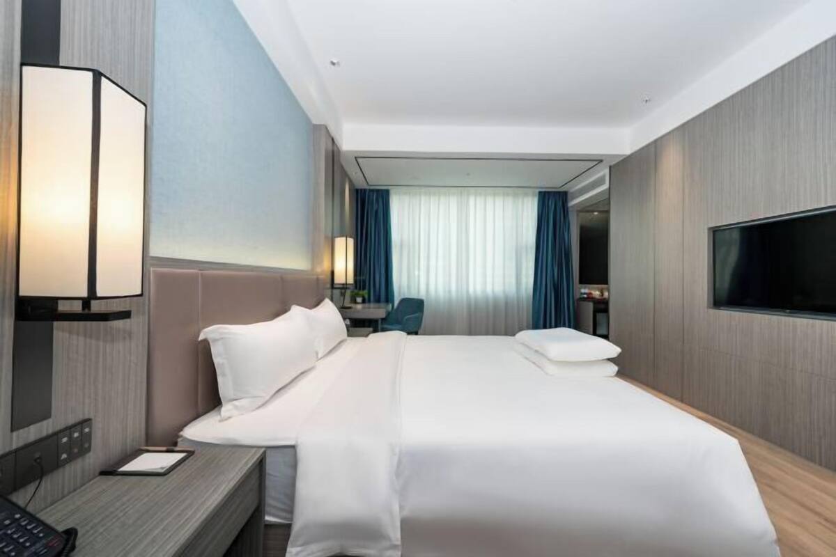 A hotel room with a bed, bedside table, lamp, TV, and a view of the corridor.