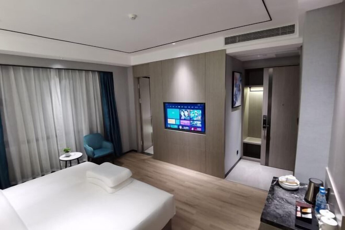 Quarto de hotel moderno com cama, televisão na parede, uma pequena mesa com uma planta e uma cadeira azul.