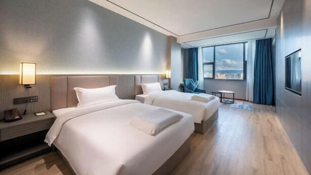 Habitación de hotel con dos camas, un escritorio, una silla, un televisor y un ventanal grande con cortinas.