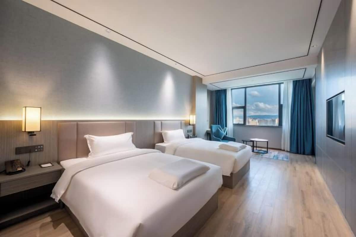A hotel room with two beds, a desk, a chair, a TV, and a large window with curtains.