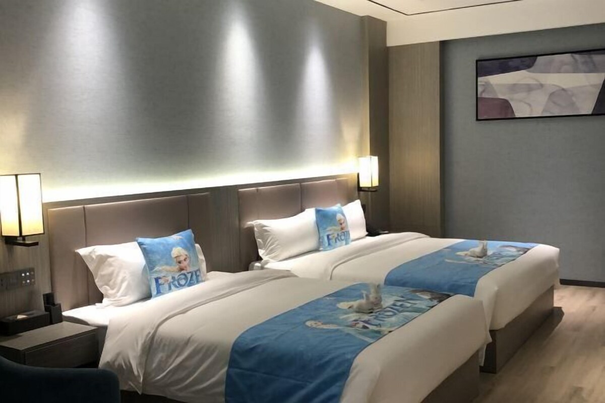 Quarto de hotel com duas camas, cada uma com travesseiro temático de Frozen, uma mesa de cabeceira com abajur e uma obra de arte pendurada na parede.