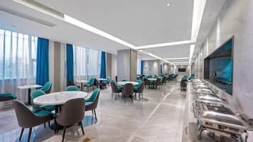 Un comedor de hotel con varias mesas y sillas, una larga barra de buffet y amplias ventanas.